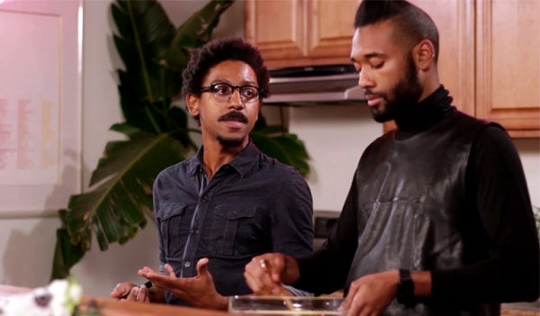 Still of Aymar Jean Christian and Elijah McKinnon during Episode 1 of "Two Queens in a Kitchen". They are cooking and engaged in a conversation in the middle of the kitchen. Directly behind them are wooden cupboards, and to their back right is the stove and a palm tree