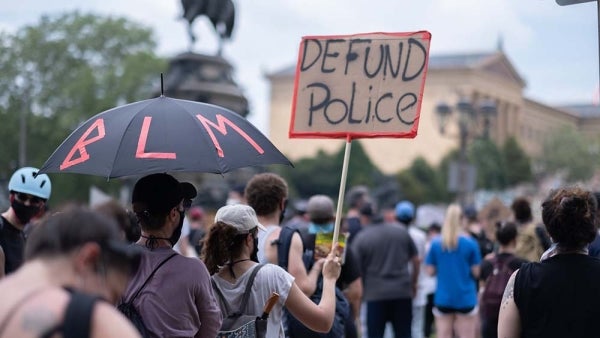 Crowd of protestors, one of whom carries an umbrella with BLM taped onto it. Another carries the sign "Defund Police"