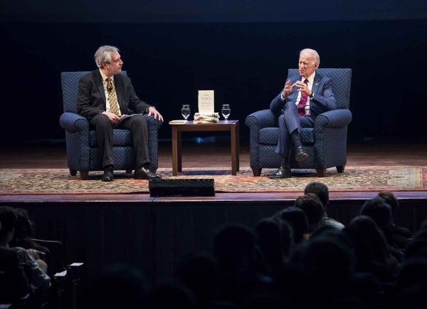 Michael Delli Carpini and Joe Biden spotlit on stage sitting in chairs against a black backdrop
