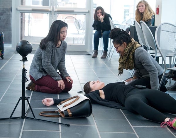 A person lies lifeless on the floor of a building as two people crouch over her and others look on from behind. There is a VR camera next to them.