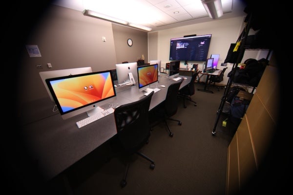 The inside of the Media Lab. Six iMac desktop computers are on a long conference table. A large TV screen is on the wall.