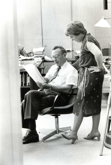 George Gerbner seated in his office with a woman standing over his shoulder