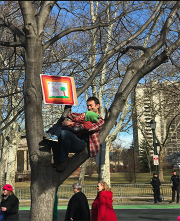 Emile Bruneau sitting in a tree holding his son and a sign that says "Respect for All"