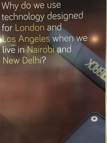 Blurred brown background with two pieces of technology of the color back to the right of the image's foreground. At the top-right, it reads "Why do we use technology designed for London and Los Angeles when we live in Nairobi and New Delhi?"