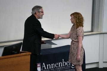 Rosemary Clark-Parsons receiving her award from a man. They are shaking hands and the man holds the certificate in front of a table with a blue cloth that has the Annenberg School for Communication logo on it. 