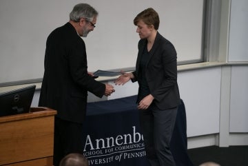 Rachel N. Stonecipher receives award from a man in front of a table with a navy blue cloth over it that has the Annenberg School for Communication logo at the front. The man holds the certificate in his left hand as he and Stonechipher are about to shake hands.
