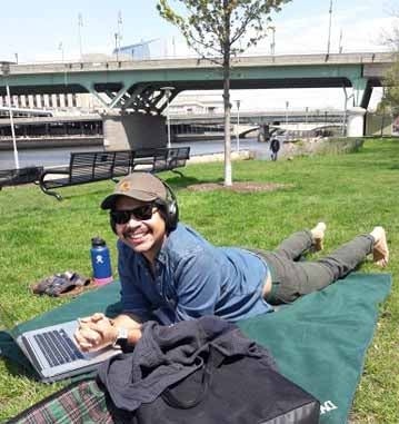 Person lying in the grass with a laptop by the side of a river with a bridge in the background