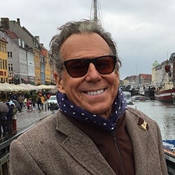 Headshot of Bill Bogs smiling for the picture on a river wearing sunglasses, a navy blue scarf with white polka dots, and light brown jacket 