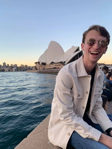 Justin Cohen sitting on a ledge with water and the Sydney Opera House behind him